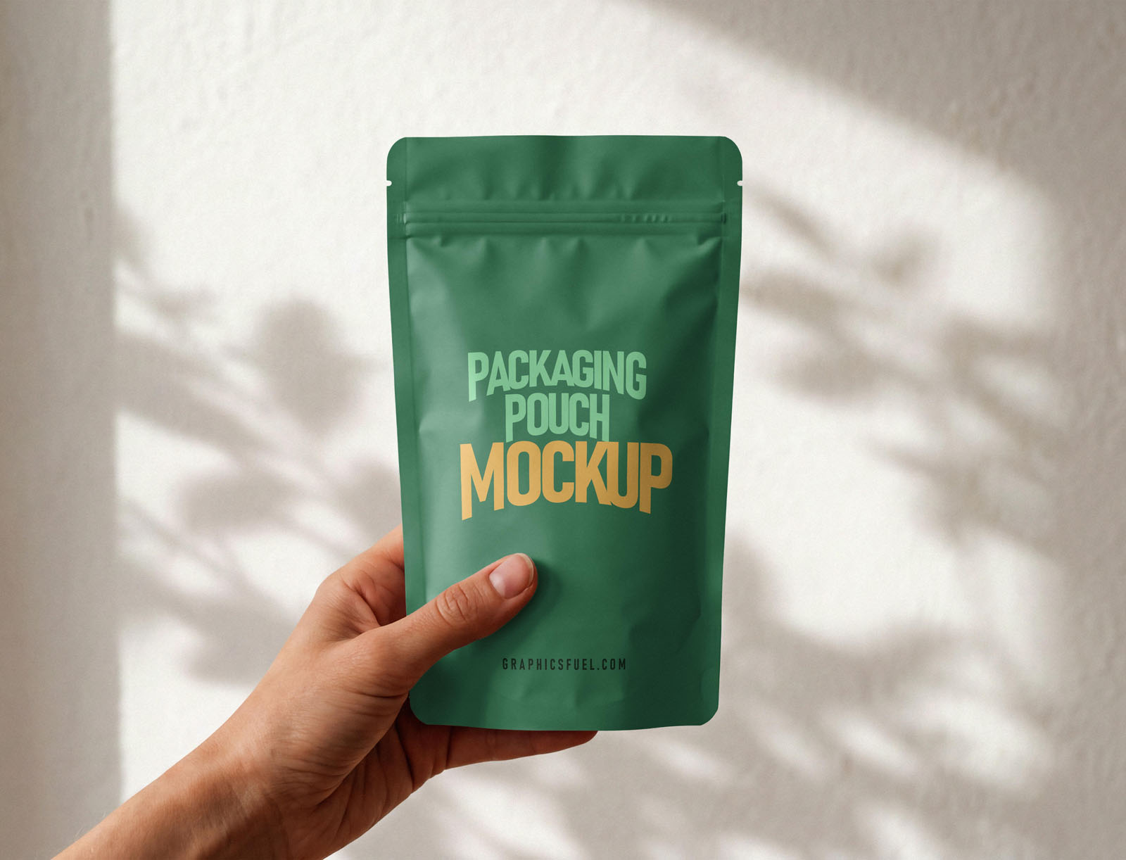 Hand holding a green packaging pouch mockup against a sunlit wall with leaf shadows
