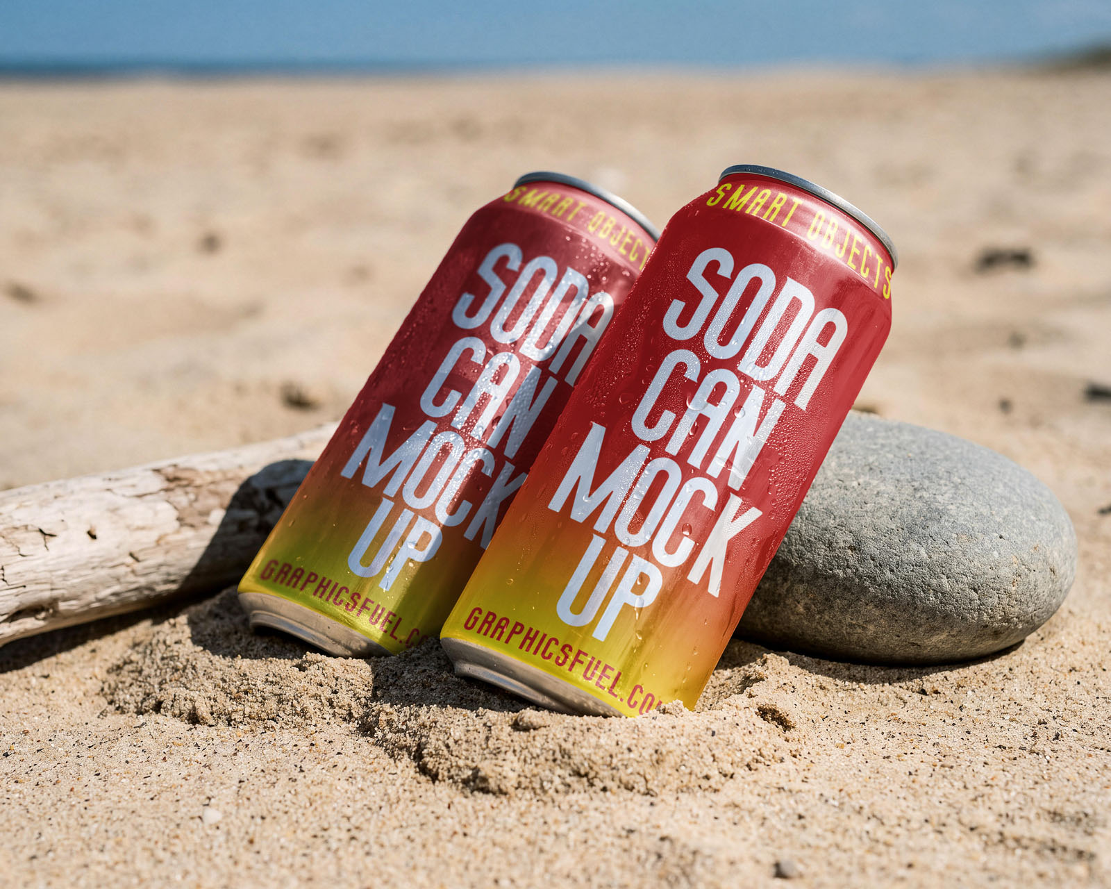 Two soda can mockups propped against driftwood and stone on sand