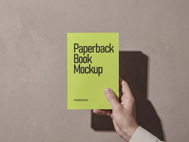 Free paperback book mockup held in hand against a warm plaster wall